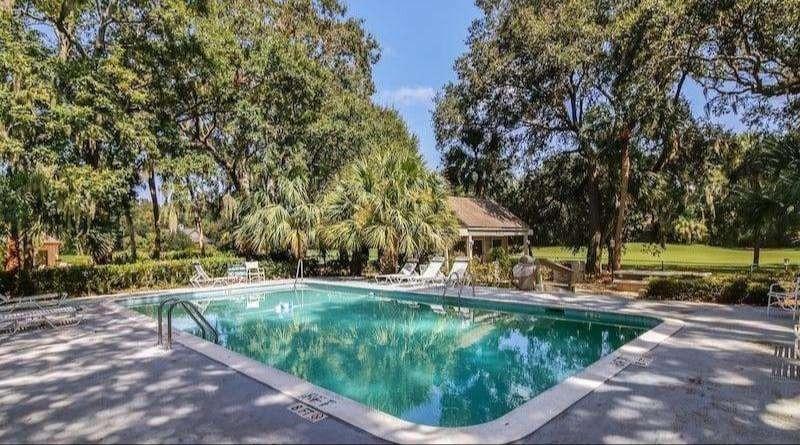An outdoor community swimming pool surrounded by a fence and lounge chairs, set against a backdrop of trees and a golf course fairway.