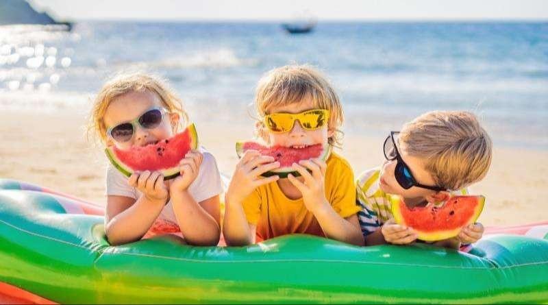 Three children eating watermelon slices on an inflatable raft at the beach