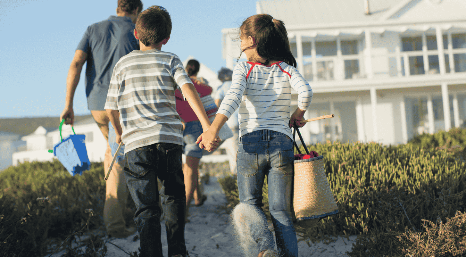 A family of four walking on a sandy path towards the beach with a rental home in the background.