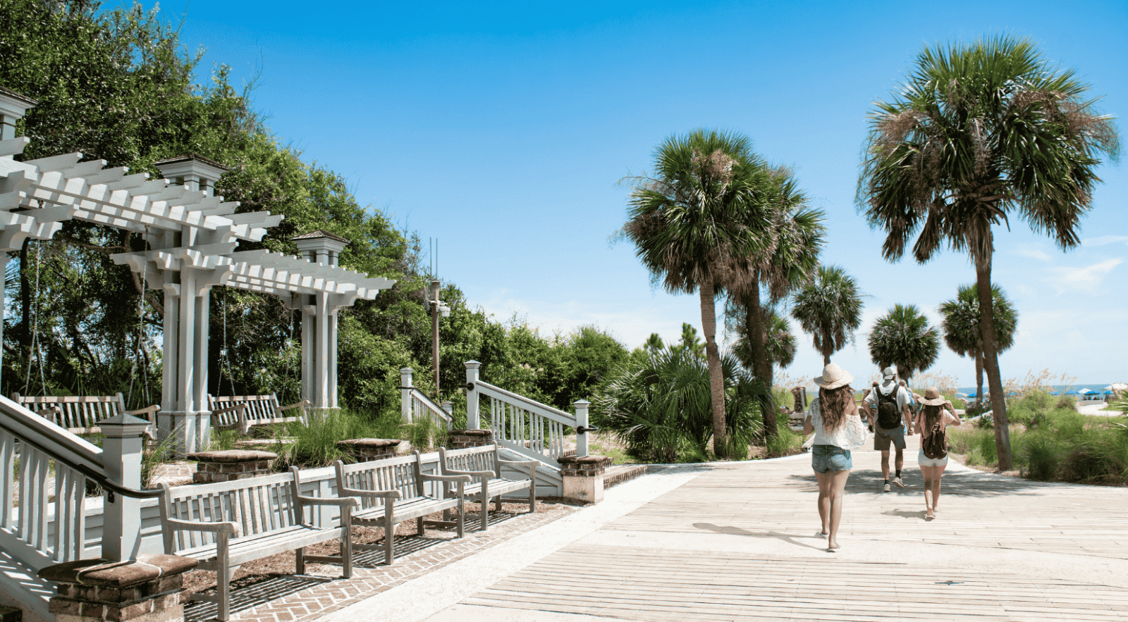 Tourists walking on a wooden boardwalk at a beach park with palm trees and white benches.